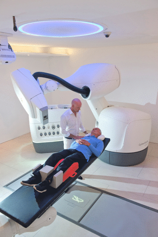 Ein Krebs-Patient im Behandlungsraum kurz vor der Radiochirurgie-Behandlung mit dem modernsten Cyberknife-System M6. Der am Cyberknife-Kopf befestigte Aufsatz zeigt den neuesten Linsenaufsatz „InCise 2 Multileaf Collimator“, der zur Patientenbehandlung weltweit erstmalig in München eingesetzt wurde. (Foto: Europäisches Cyberknife Zentrum München-Großhadern) / A cancer patient in the treatment room just before radiosurgery treatment using the most advanced CyberKnife System M6. The CyberKnife headmounted attachment shows the latest lens attachment “InCise 2 Multileaf Collimator”, which was used for patient care for the first time worldwide in Munich. 
(Photo: European Cyberknife Center Munich-Grosshadern) 