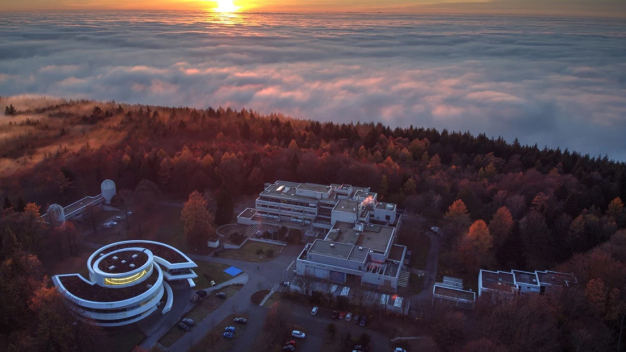 Luftbild des MPIA-Campus auf dem Königstuhl mit dem spiralförmigen Haus der Astronomie (links unten), den Teleskop-Kuppelgebäuden des Elsässer-Labors (links) und dem MPIA-Hauptgebäude (Mitte). Der Erweiterungsbau wird sich in Richtung der Kuppelgebäude an das Südende des Hauptgebäudes anschließen. (Quelle: Domink Elsässer) / Aerial view of the MPIA campus on the Königstuhl with the spiral-shaped House of Astronomy (down left), the telescope dome buildings of the Elsässer Laboratory (left) and the MPIA main building (center). The extension building will adjoin the south end of the main building in the direction of the domed buildings. (Credit: Domink Elsässer) Luftbild des MPIA-Campus auf dem Königstuhl mit dem spiralförmigen Haus der Astronomie (links unten), den Teleskop-Kuppelgebäuden des Elsässer-Labors (links) und dem MPIA-Hauptgebäude (Mitte). Der Erweiterungsbau wird sich in Richtung der Kuppelgebäude an das Südende des Hauptgebäudes anschließen. (Quelle: Domink Elsässer) / Aerial view of the MPIA campus on the Königstuhl with the spiral-shaped House of Astronomy (down left), the telescope dome buildings of the Elsässer Laboratory (left) and the MPIA main building (center). The extension building will adjoin the south end of the main building in the direction of the domed buildings. (Credit: Domink Elsässer)